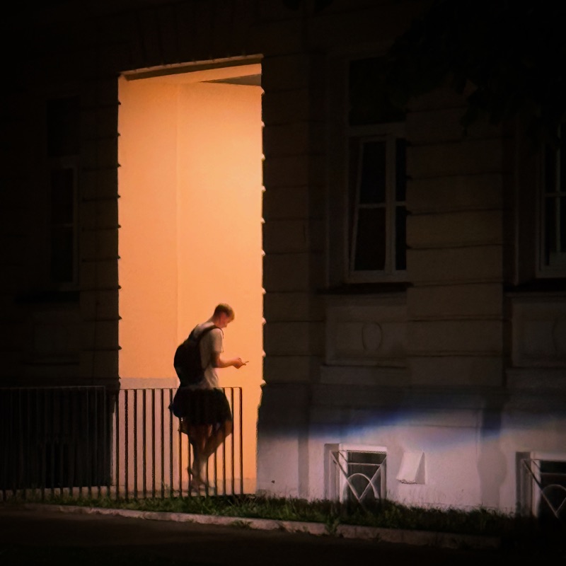A boy with a backpack looking at his phone at night, silhouetted against a warm light on a building wall
